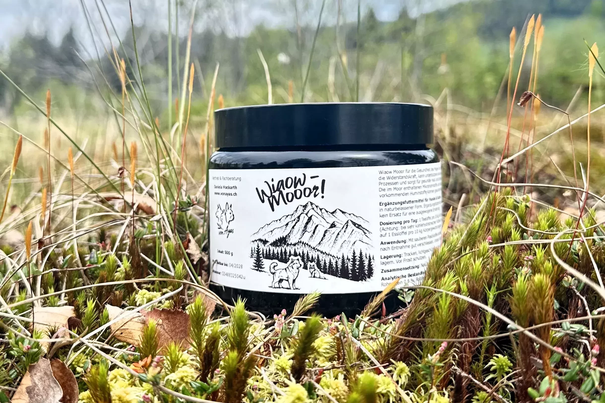 Wiaow Mooor Naturmoor im schwarzen Glastiegel steht draussen zwischen Moos und Gras.